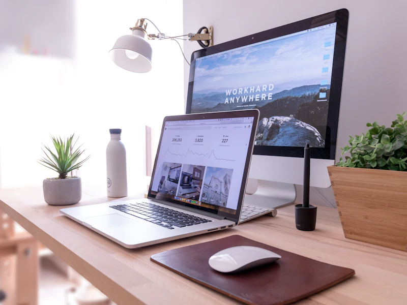 A tidy desk by a window with a laptop open in front of a larger monitor.