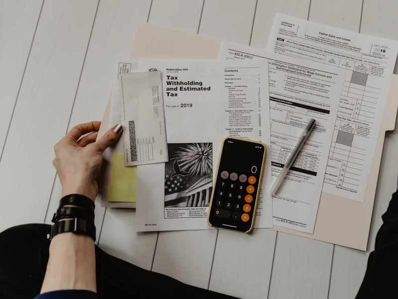 Tax forms and statements spread across a white desk beside a calculator app on a phone.