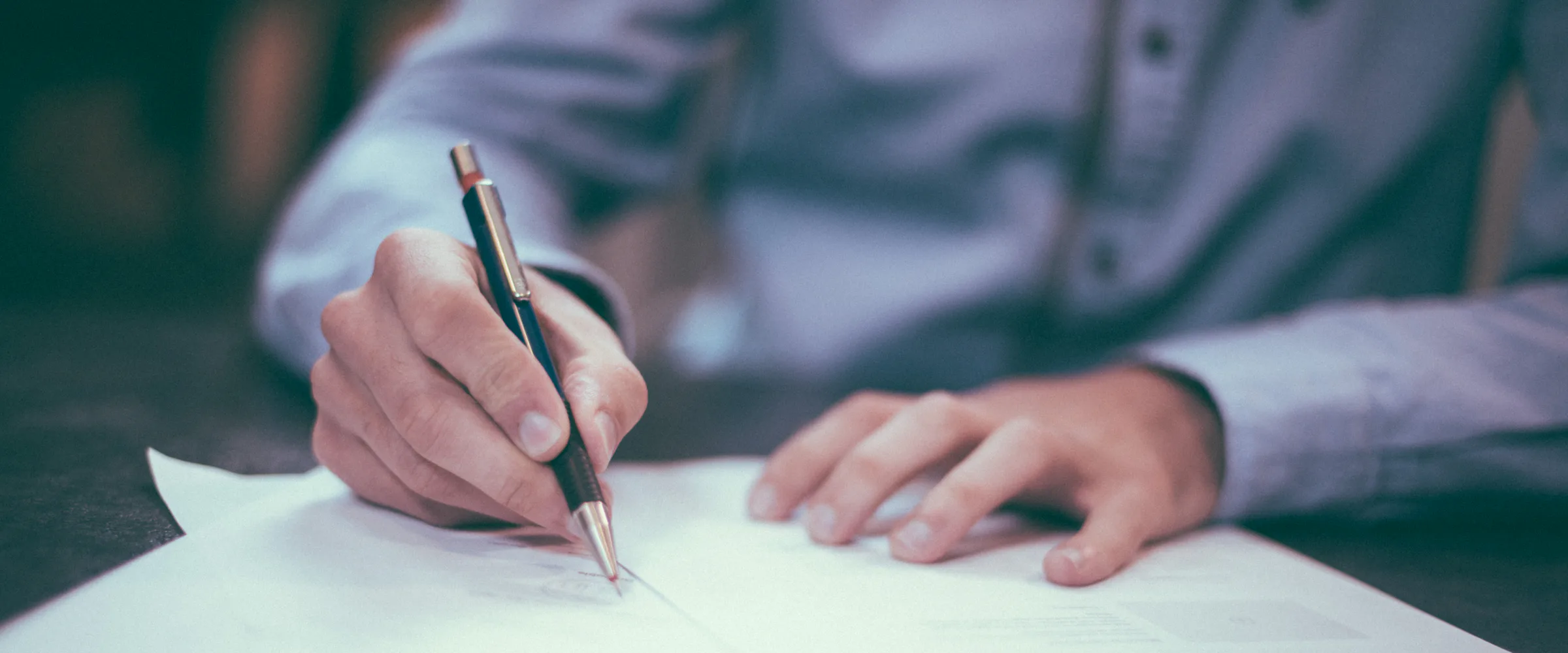 A hand signing a printed document at a desk, with a second page held flat beside it.